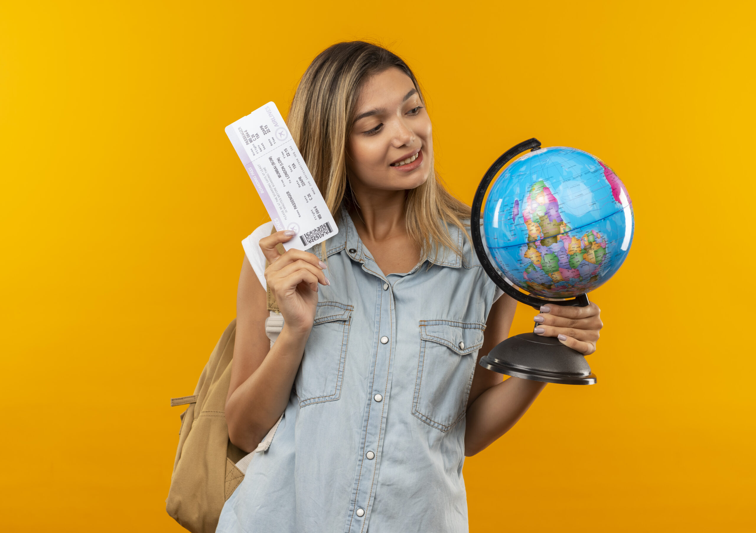 pleased young pretty student girl wearing back bag holding globe and airplane ticket looking at globe isolated on orange background